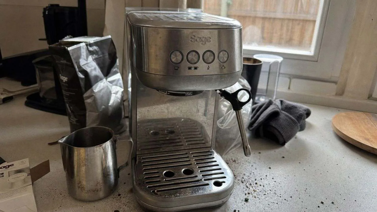 A compact stainless espresso machine at the back of a kitchen worktop, with grounds scattered around its base, a milk jug and an open bag of beans beside it.