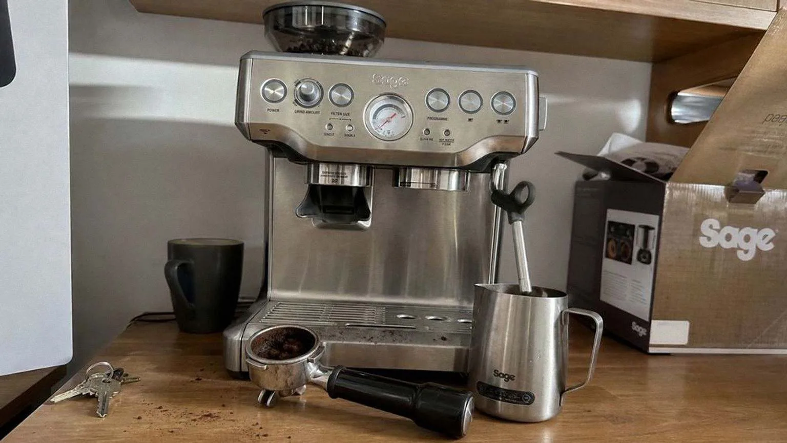 The Barista Express on a domestic worktop beside the wall cupboard it has to fit under, with a used portafilter and a milk jug at its side.